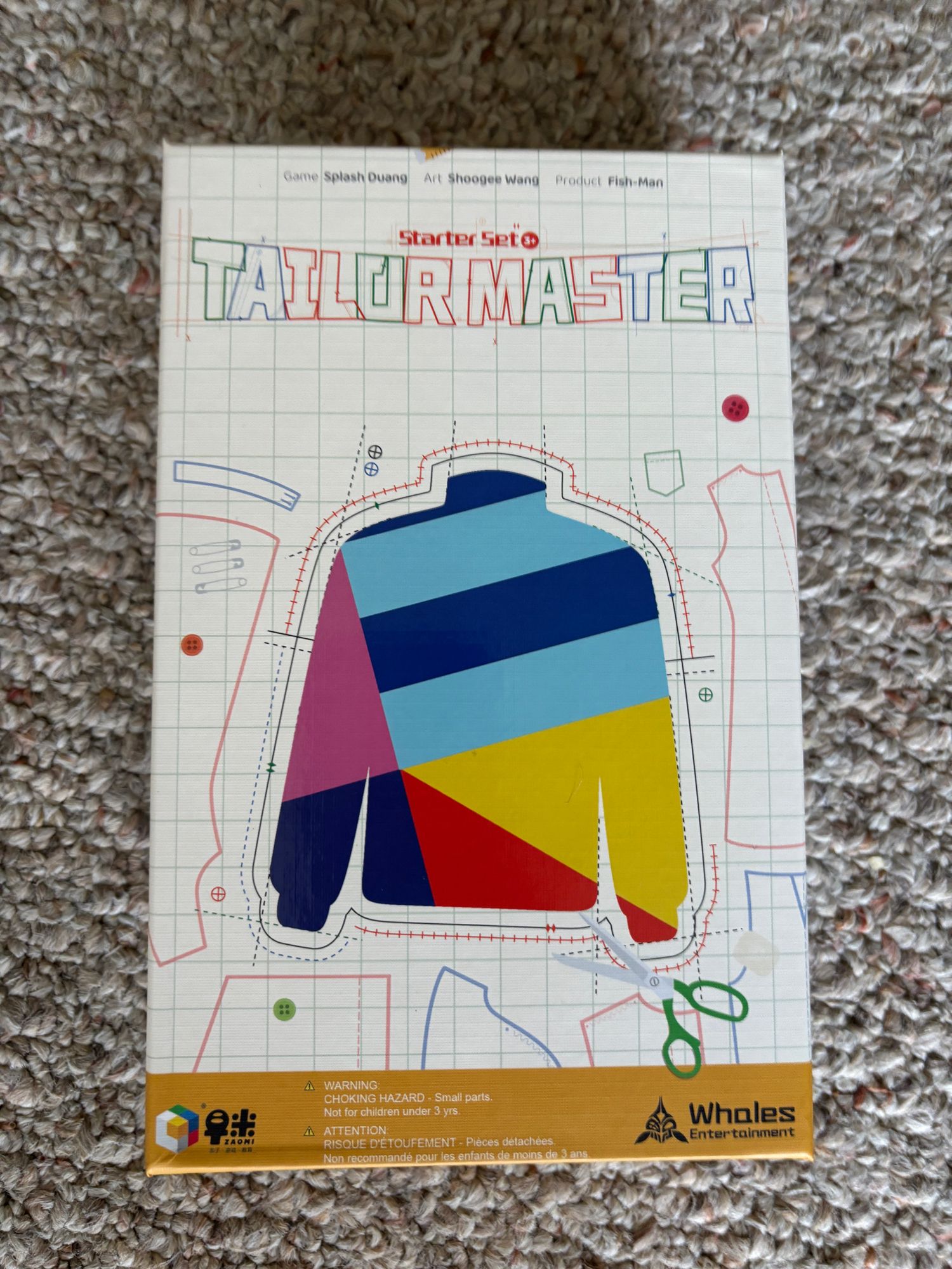Box cover for the board game Tailor Master Starter Set by Whales Entertainment, featuring a colorful sweater pattern design with scissors and sewing line details.